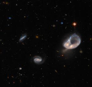 Two galaxies right of center form a ring shape. The ring is narrow and blue, and the cores of the two galaxies form a bulge on the ring’s side. A bright, orange star lies above the ring. Two smaller spiral galaxies appear left of center, as well as a few stars. The background is black and speckled with very small stars and galaxies.
